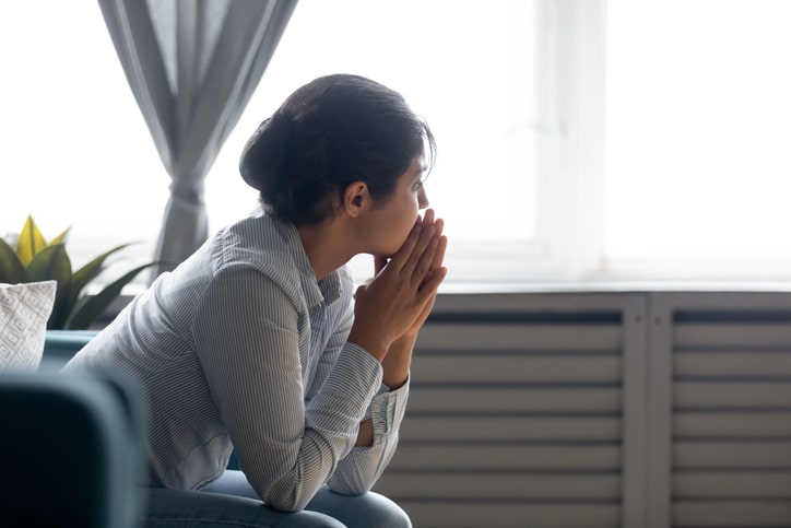 A young woman stares pensively out the window with her hands to her chin after losing a loved one