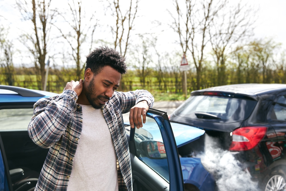 A man leans against the passenger side door in the aftermath of an accident and holds his hand to his neck in pain