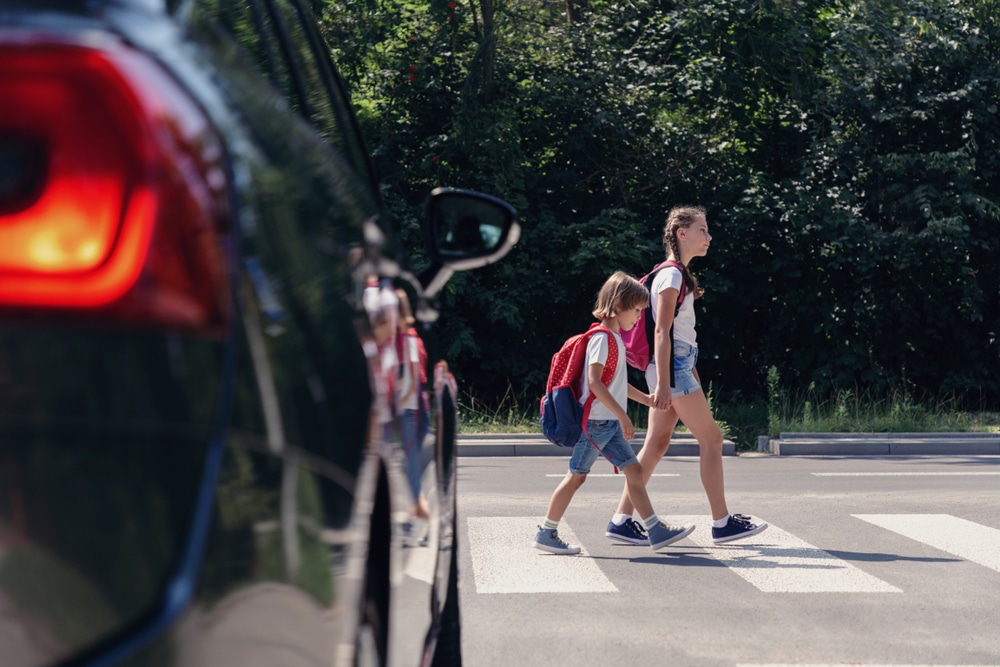 Tow young children with backpacks cross at a marked crosswalk with a stopped car in front of them