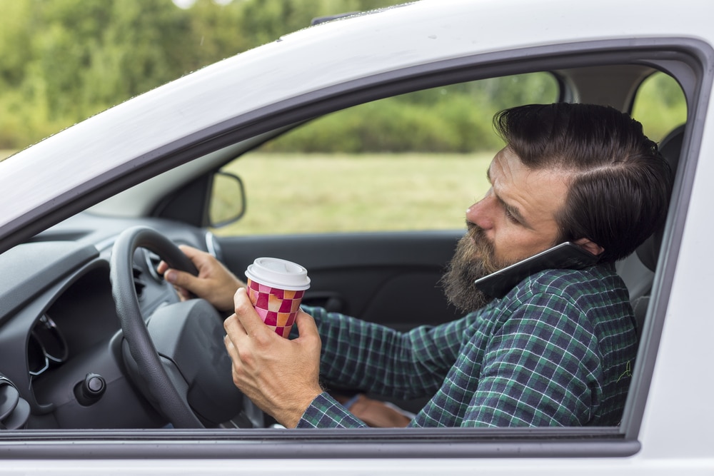 A man holds a coffee and has his phone held up by his shoulder as he tries to drive