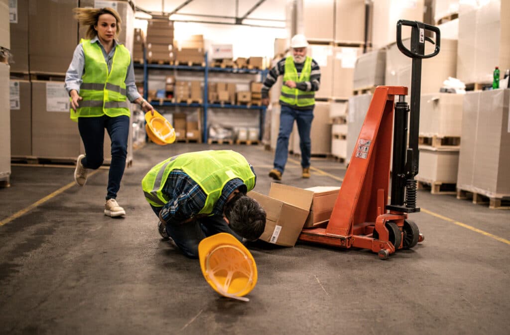 Man in a Hi-Vis safety vest bent over in pain from a work injury in a factory, with other workers around him.